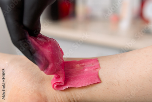 Close-up. Woman cosmetologist in black gloves applies pink wax or sugar paste on client's legs before depilation, epilation