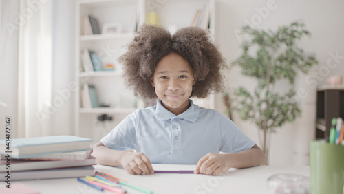 Adorable kid with natural black hair smiling at camera, sitting at table with pens and books, ready for studying