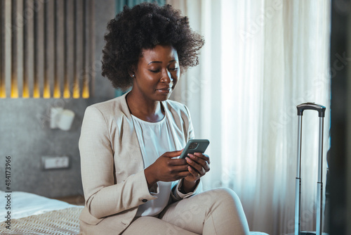 Young black businesswoman just arrived at the hotel taking a rest from a trip, using a smartphone. She working from hotel room on business trip, using smart phone. 