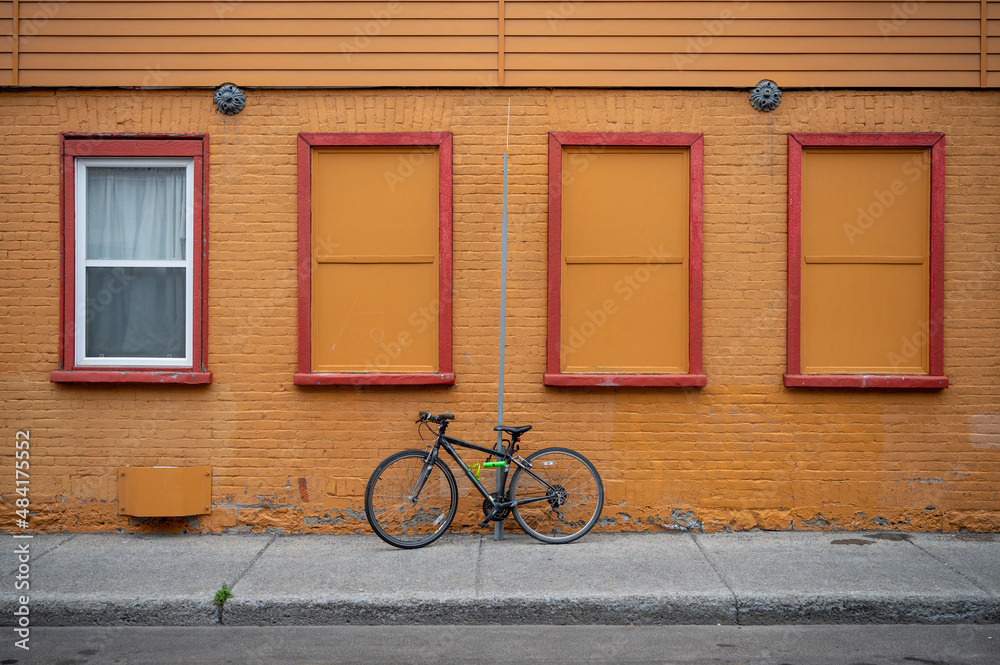 A Bicycle Rests Locked Up To A Thin Post With A Yellow Brick Building With Four Windows Behind It. 