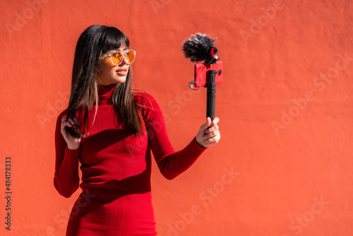 Wallpaper Mural Brunette girl recording a video blog with the mobile phone and the microphone, wearing a dress for social networks. Caucasian pretty a red background, copy space and paste Torontodigital.ca