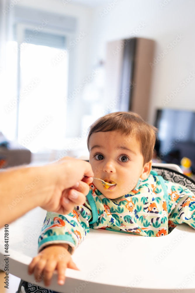 Feeding his seven-month-old baby at home, his first vegetable purees, first-person view of the father spooning pumpkin, carrot and potato puree into his mouth Feeding his seven-month-old baby at home, his first vegetable purees, first-person view of the father spooning pumpkin, carrot and potato puree into his mouth