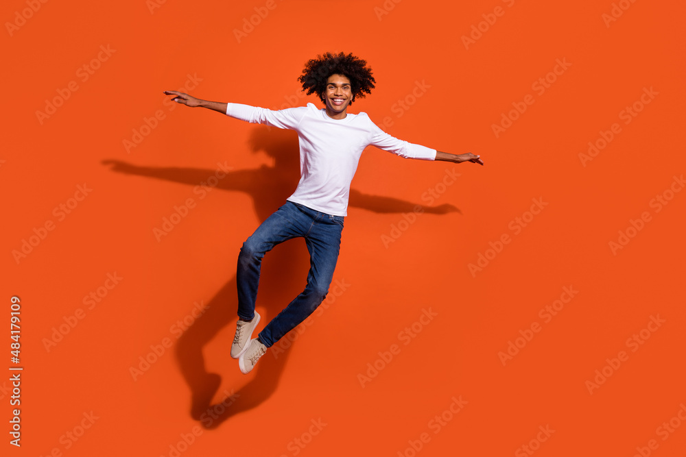 Photo of excited cute guy wear white shirt jumping high arms sides empty space isolated orange color background Photo of excited cute guy wear white shirt jumping high arms sides empty space isolated orange color background