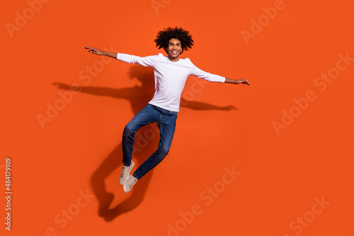 Photo of excited cute guy wear white shirt jumping high arms sides empty space isolated orange color background