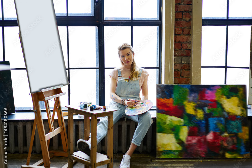 Portrait of attractive female mastery with pallet resting at studio windowsill and looking at camera during weekend daytime for creating paintings, Caucasian woman posing near easel with canvas Portrait of attractive female mastery with pallet resting at studio windowsill and looking at camera during weekend daytime for creating paintings, Caucasian woman posing near easel with canvas