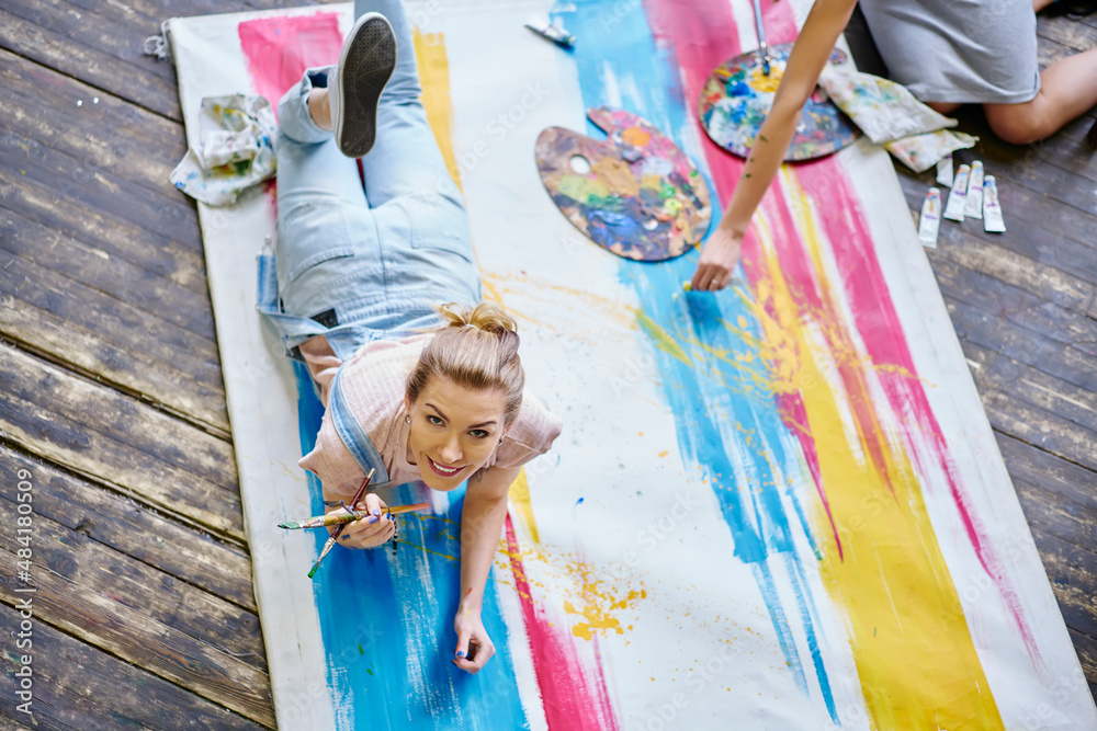 Portrait of cheerful female artist dressed in jeans wear smiling at camera while lying at colored canvas enjoying leisure in art studio, happy Caucasian woman with paintbrush posing indoors