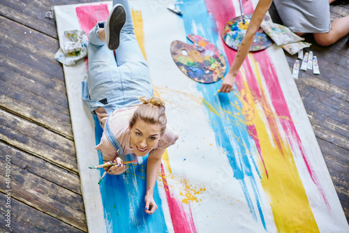 Portrait of cheerful female artist dressed in jeans wear smiling at camera while lying at colored canvas enjoying leisure in art studio, happy Caucasian woman with paintbrush posing indoors © BullRun