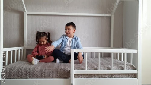 Two Children brother and sister are sitting in a Montessori bed and playing Video games. Girl is holding a gamepad and the boy is looking to her.
