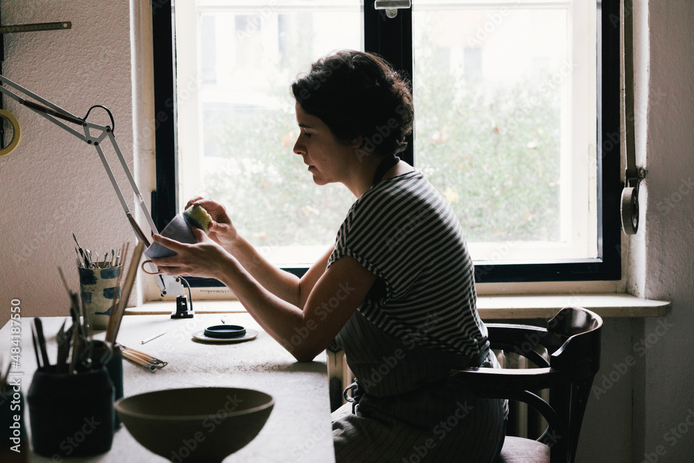 Mature craftswoman painting cup with cleaning sponge while sitting on chair Mature craftswoman painting cup with cleaning sponge while sitting on chair
