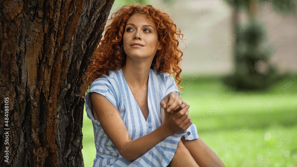 pleased redhead woman in blue striped dress sitting under tree trunk. pleased redhead woman in blue striped dress sitting under tree trunk.