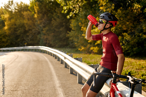 Fototapeta Naklejka Na Ścianę i Meble -  Happy smiling cyclist man drinking water while resting after intensive driving a bike, young caucasian male cyclist sitting near road, after sunny morning workout, healthy, sport lifestyle