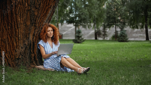 full length of curly redhead woman in dress listening music while using laptop in park.