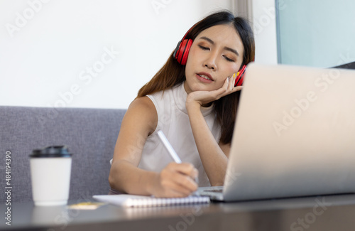 Asian woman working on laptop at home having an online meeting and writing notes in a notepad.