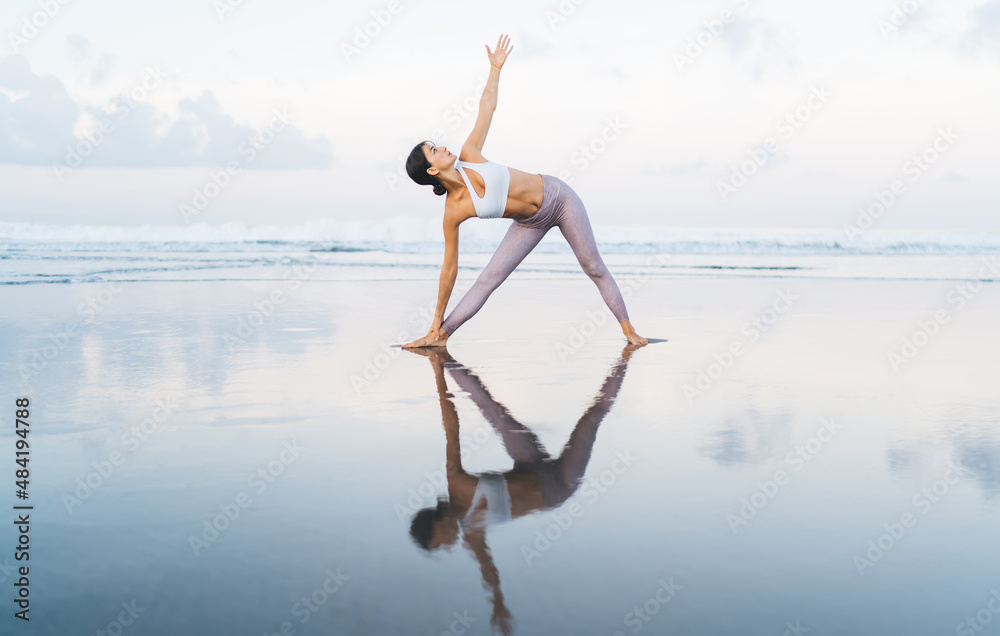 Fototapeta premium Caucasian female in sportswear enjoying morning time for holistic healing during yoga meditation at seashore, slim woman stretching body muscles during concentrated pilates workout at coastline