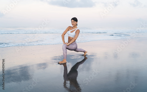 Portrait of young Caucaisan woman in sportive clothes looking at camera during morning stretching workout at coastline beach  attractive fit girl slimming while doing pilates and aerobic exercises