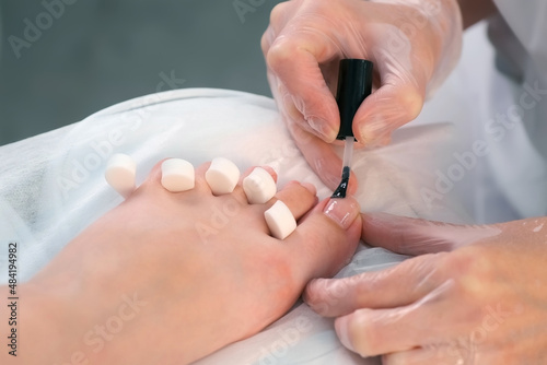 Pedicurist master is applying transparent base coal for shellac on woman's nails on toes, closeup hands. Preparing nails for shellac in beauty salon. Hygiene and care for feet. photo