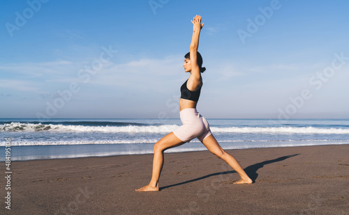 Side view of Caucasian female in active wear doing exercises at coastline beach recreating and keeping perfect body shape  flexible woman have morning yoga near ocean enjoying time for meditation