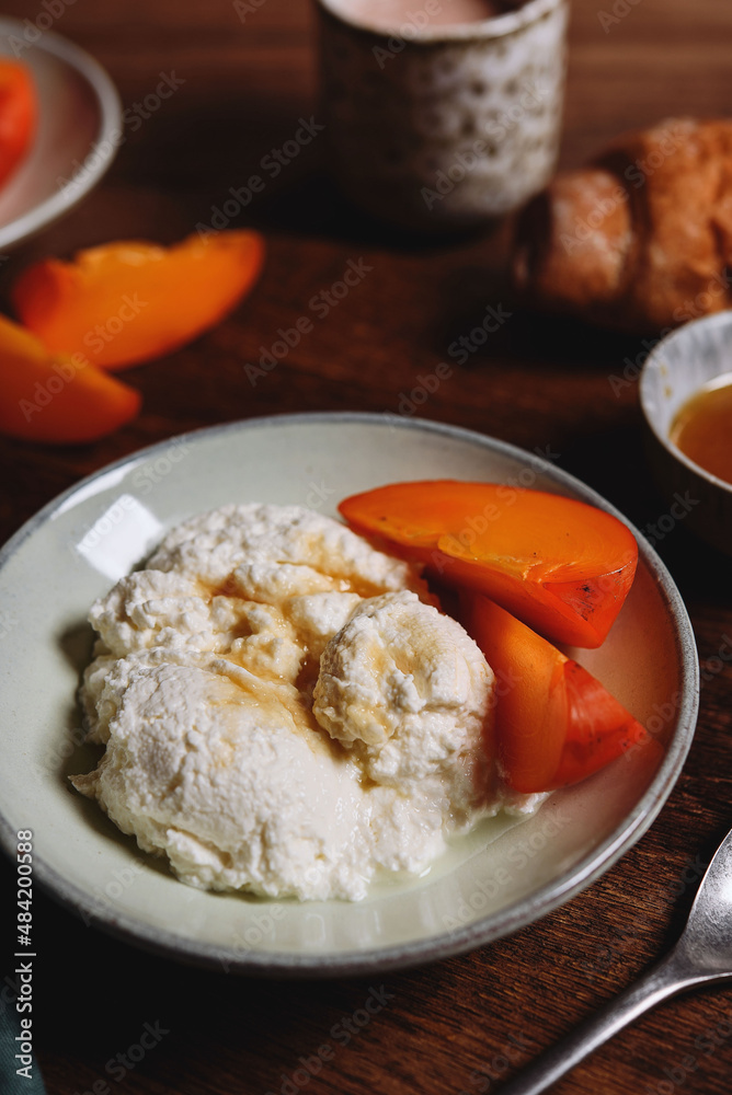 Homemade ricotta cheese or cheese curd served with fresh persimmon and honey and croissante on wooden background. Healthy breakfast. Haze effect. Selective focus Homemade ricotta cheese or cheese curd served with fresh persimmon and honey and croissante on wooden background. Healthy breakfast. Haze effect. Selective focus
