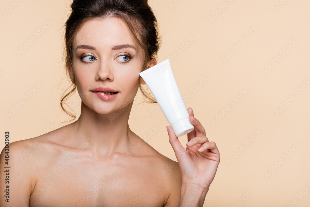Pensive model with naked shoulders holding cosmetic lotion isolated on beige.