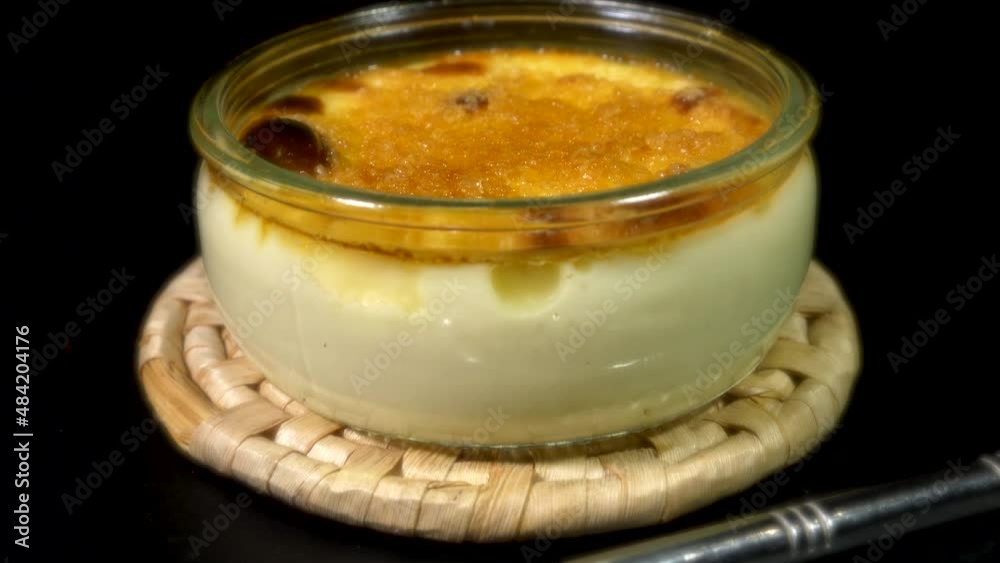 Closeup forward dolly shot of a bubbling hot Crème Brûlée – a custard base dessert, topped with caramelized brown sugar, ready to eat from a shallow glass jar with the spoon in the foreground.