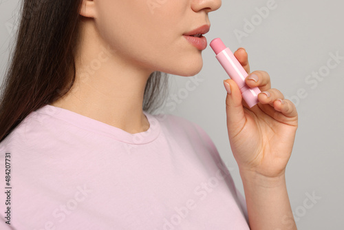 Young woman applying lip balm on grey background, closeup