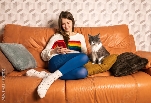 Young cute girl in glasses wearing white sweater with rainbow art and tight blue leggins sitting on orange leather sofa at home with her gray female cat working remotely with smartphone © Andrii