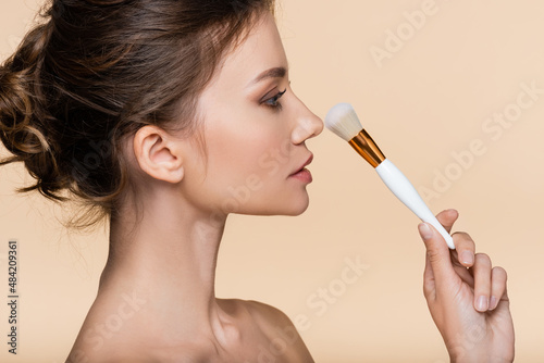 Side view of brunette woman holding cosmetic brush near nose isolated on beige.