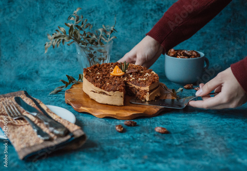 Women's hands take a piece of homemade cake with chocolate and cream with a spatula next to a plate with a fork and knife and a mug filled with nuts on dark background. Home baking