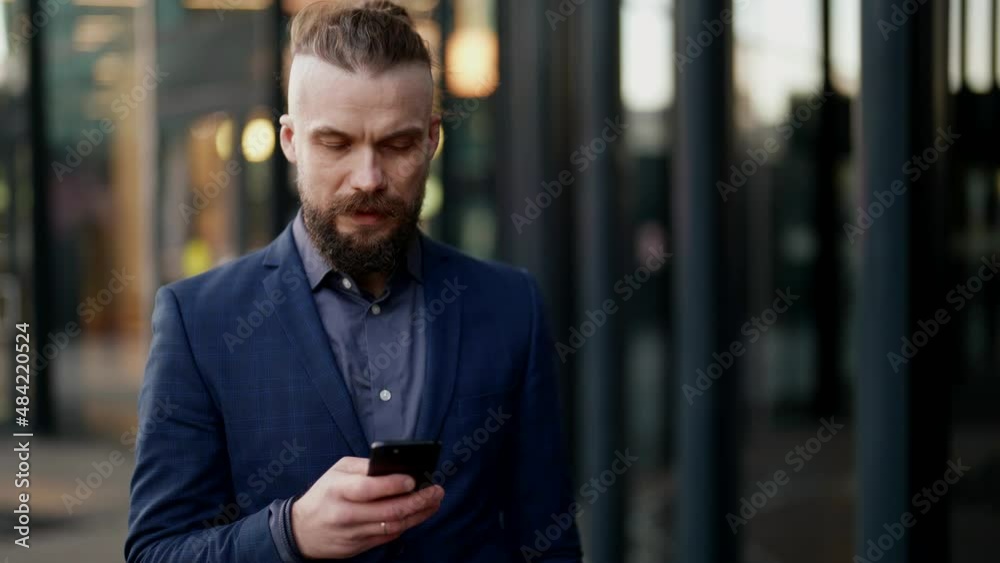 a mustachioed and bearded man in a plaid jacket looks discontentedly at the phone screen and walks along a blurry modern building a mustachioed and bearded man in a plaid jacket looks discontentedly at the phone screen and walks along a blurry modern building