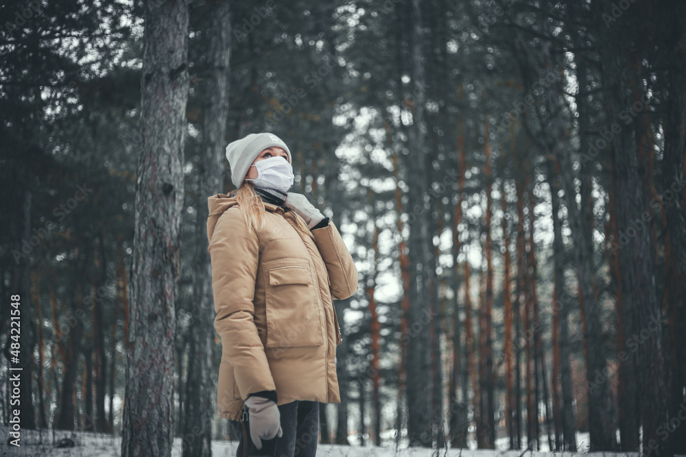 A young woman with blond hair in a medical mask walks in the winter forest.