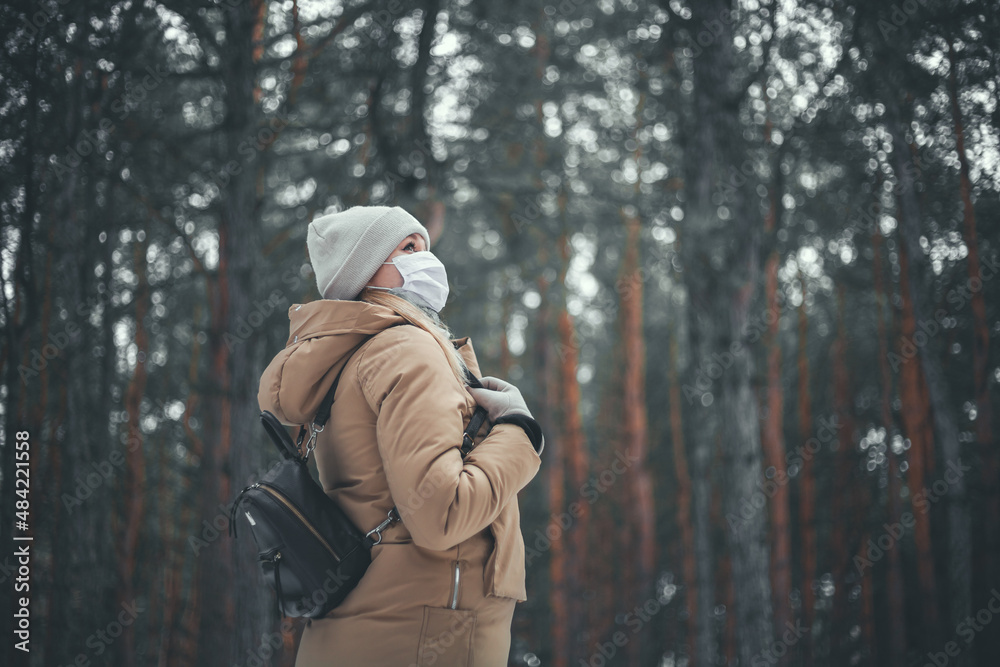 A young girl with blond hair in a medical mask walks in the winter forest.