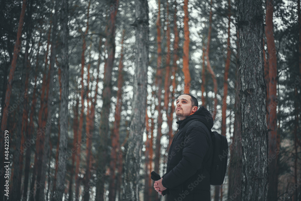 A young attractive man with short hair in a black winter coat poses against the backdrop of a winter forest.