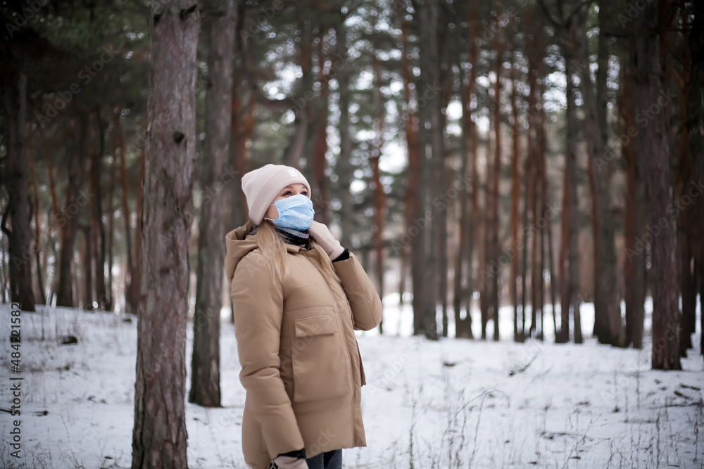 A young woman in winter clothes and a medical mask is walking in the forest.