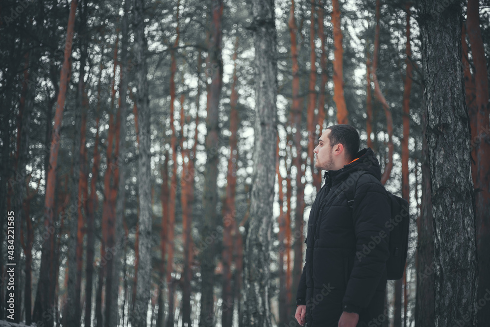 A young attractive man with a short beard and short hair in a black winter coat poses against the backdrop of a winter forest.