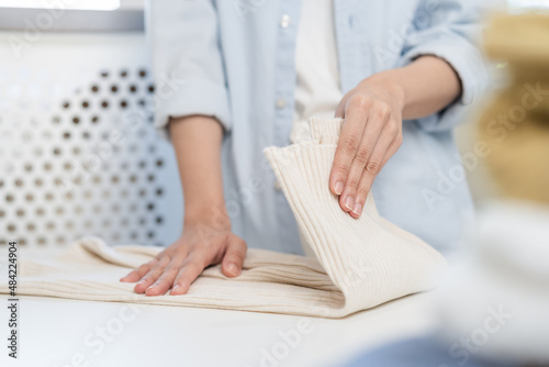 Housewife, asian young woman hand in many folding freshly shirts, sweaters or dress on desk, table after washing clean clothes and drying, making household working in room at home. Laundry and maid.