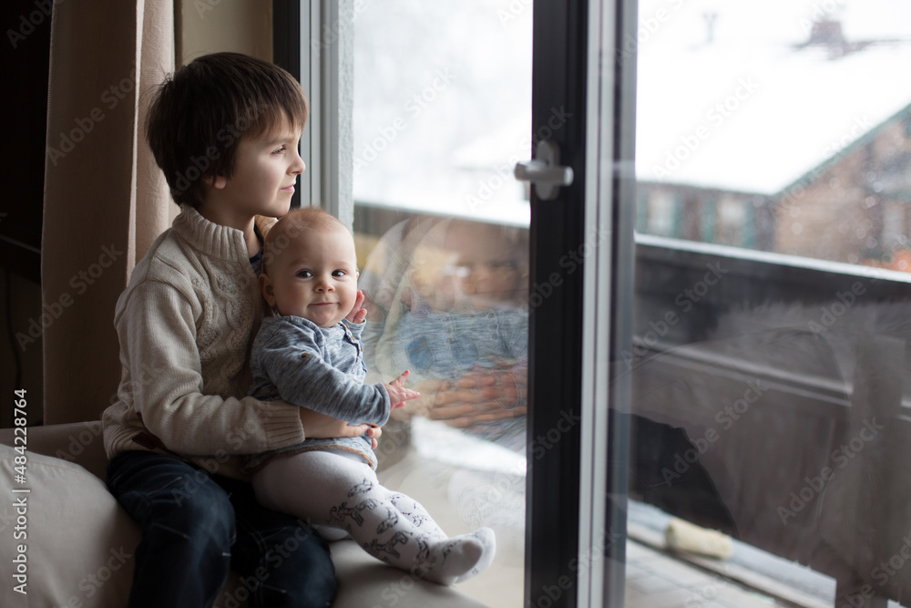 Preschool boy, holding his baby brother, sitting by the window in living room, looking at a snowy landscape outdoors Preschool boy, holding his baby brother, sitting by the window in living room, looking at a snowy landscape outdoors