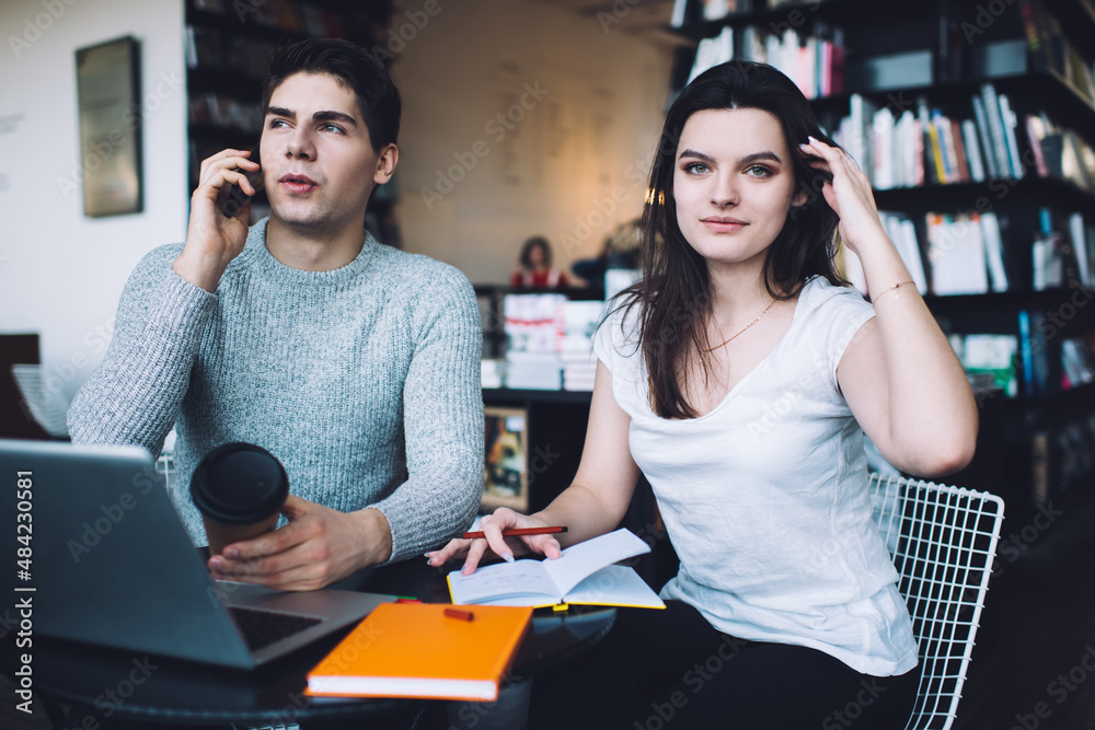 Concentrated young man chatting on smartphone sitting in workspace with colleague Concentrated young man chatting on smartphone sitting in workspace with colleague