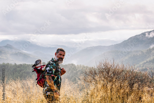 Front view of traveler looking at camera with his dog in backpack in the mountains