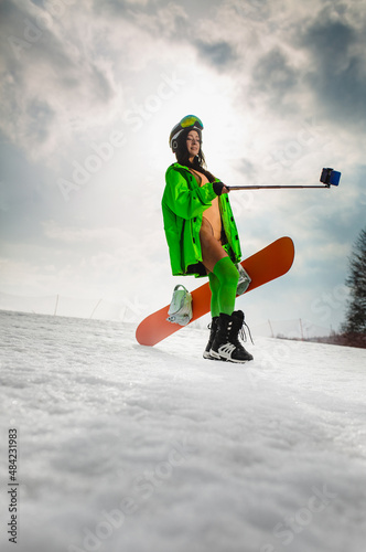 Young beautiful woman in an erotic suit using smartphone on a stick to making selfie with a snowboard on a ski slope