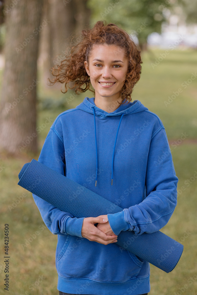 a European woman in a blue hoodie with a blue yoga mat chooses a place to do sports in the park. outdoor sports, yoga and meditation and fitness for body health. spiritual development. outdoor sports a European woman in a blue hoodie with a blue yoga mat chooses a place to do sports in the park. outdoor sports, yoga and meditation and fitness for body health. spiritual development. outdoor sports
