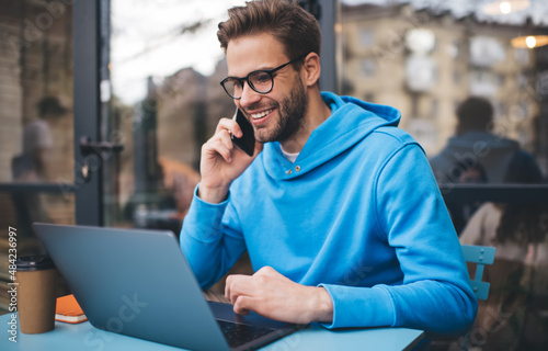 Funny hipster guy reading text publication during social networking via netbook app, happy male freelancer in eyewear doing distance job on netbook connecting to 4g for making roaming mobile calling