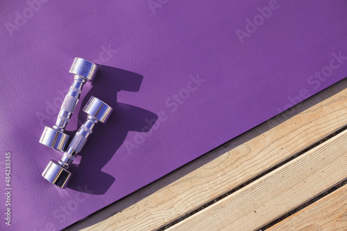 Purple yoga mat with metal chromed dumbbells. Sport fitness accessories outdoors on wooden floor on sunny day.