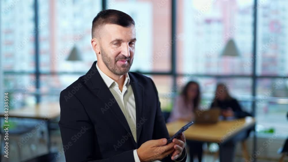 Bearded man receives a call on the phone. Businessman having conversation and gesturing with his hand. Women sitting at the desk at backdrop in blur. Bearded man receives a call on the phone. Businessman having conversation and gesturing with his hand. Women sitting at the desk at backdrop in blur.