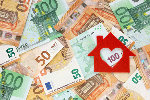 Red felt small house on right side of 50 and 100 euro banknotes background. Heart-shaped hole is cut instead of a window. Real estate loan concept. Money for a dream house. Top view. Place for text.