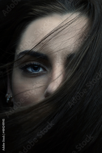 Beauty and fashion concept. Close-up beautiful woman with blue eyes portrait covering part of her face with hairs by blowing wind. Mysterious and sensual look. Eye is in camera focus