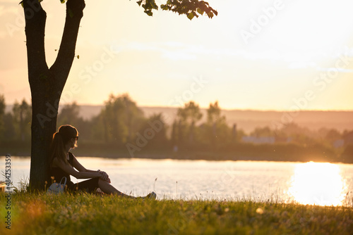 Young woman in casual outfit sitting on green grass lawn relaxing on lake side on warm evening. Summer vacations and travelling concept