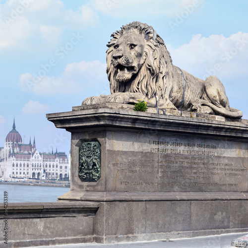 a concrete sculpture of a formidable lion on the railing of a bridge over the Danube River with the building of the Hungarian Parliament in the background