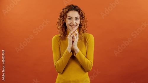 Attractive redhead caucasian girl in orange jumper twiddle her fingers and conceiving a funny plan isolated on an orange studio background.