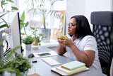 thoughtful american black lady drinking tea looking at pc computer, work day alone in office ext to window with plants. side view on confident woman. indoors. business people concept