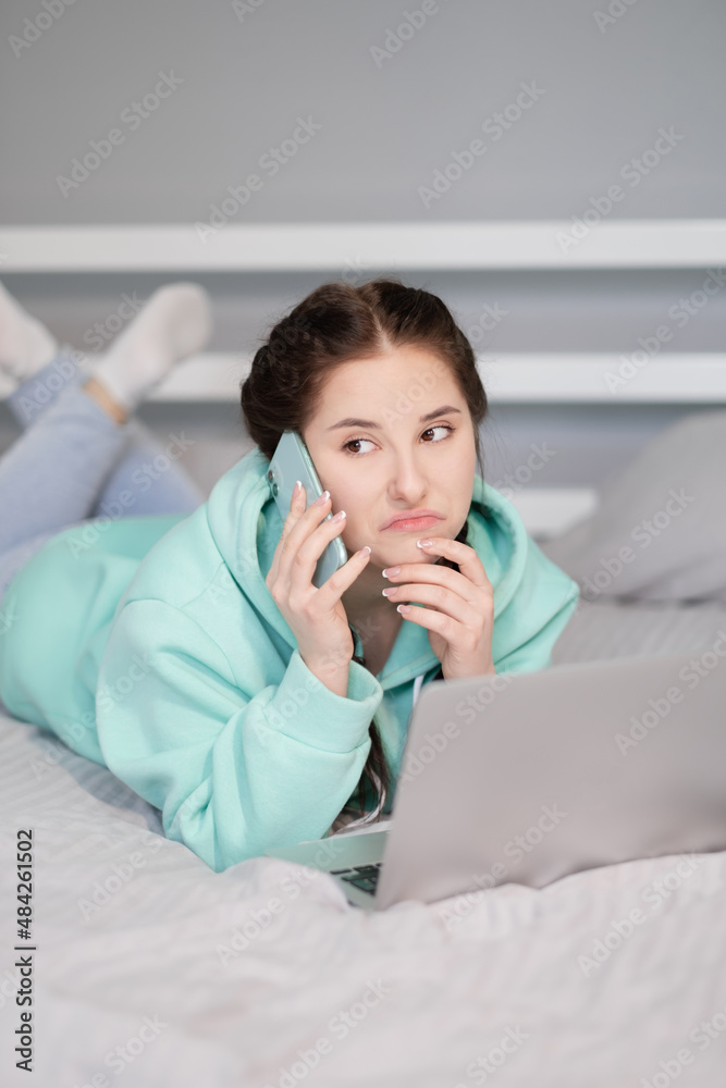 © OliaVesna - Distance learning online education and work. Happy brunette woman girl working office work remotely from home in turquoise hoodie and jeans on bed. Talking phone. Using computer laptop